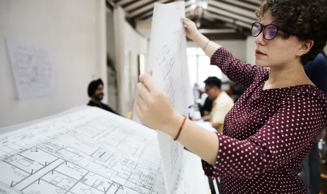 Arquitecta revisando planos en una mesa de trabajo en un estudio de arquitectura con equipo al fondo.