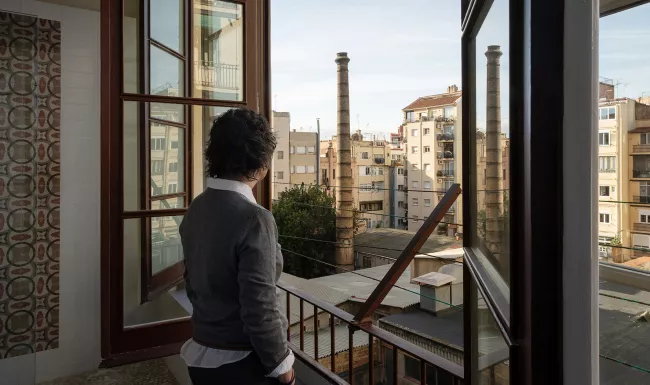 Persona d'esquena mirant per una finestra cap a l'interior d'illa de l'Eixample, amb dues xemeneies industrials de maó al fons.
