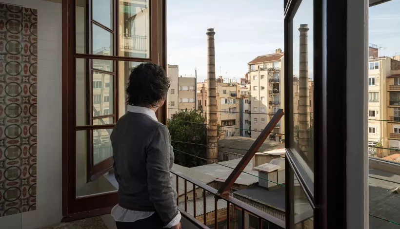 Persona d'esquena mirant per una finestra cap a l'interior d'illa de l'Eixample, amb dues xemeneies industrials de maó al fons.