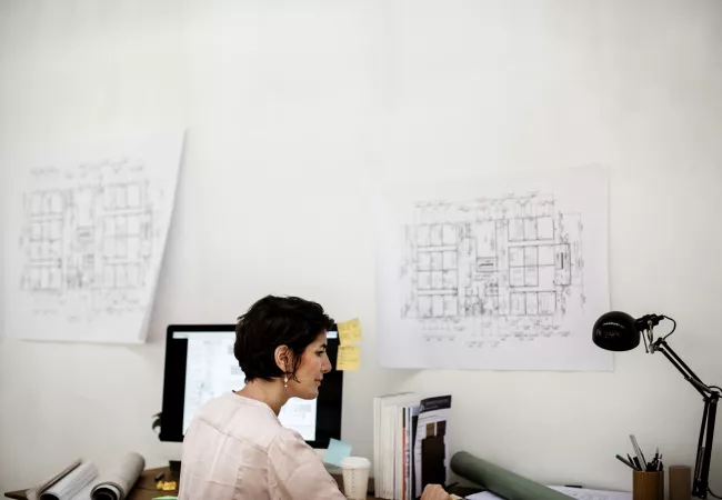 Arquitecta trabajando en su estudio frente al ordenador, con planos de planta colgados en la pared al fondo.