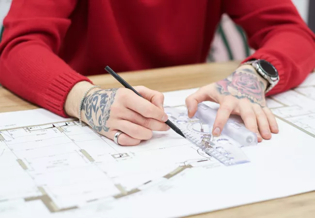 Arquitecto dibujando sobre planos con lápiz y escuadra en una mesa de trabajo.