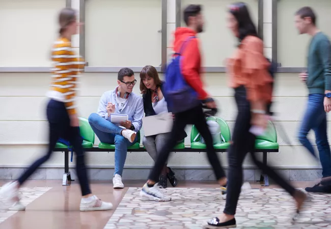 Dos estudiants consulten recursos en un portàtil en un passadís del campus mentre altres estudiants hi circulen al voltant