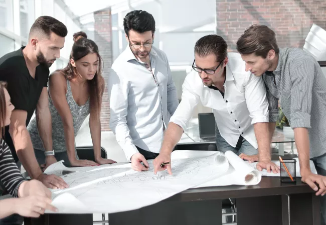 Arquitecte experimentat guia un grup d'estudiants i arquitectes novells durant una sessió formativa sobre un plànol
