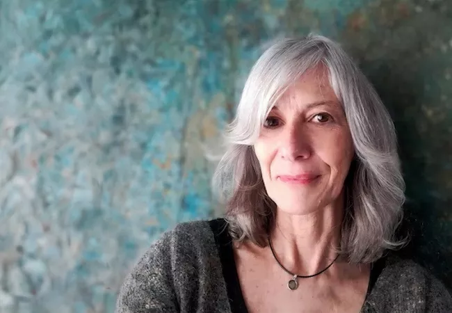 Retrato de María Teresa Alonso Argul, con el pelo canoso y jersey gris, sonriendo ante una pared de tonos turquesa